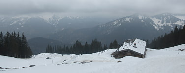 Eine schneebedeckte Berghütte am Hang vor einer wolkenverhangenen Bergkulisse