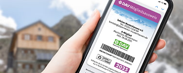 Hand mit Handy und offener DAV-App vor Berghütte 