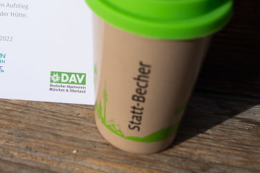 brauner Mehrweg-Kaffeebecher mit grünem Deckel und Audruck 