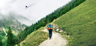 Geteiltes Bild: Im Vordergrund Sommerlandschaft mit Wanderndem am Berg. Im Hintergrund Skifahrer an verschneitem Berghang