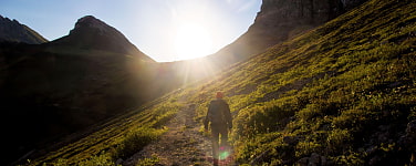 Wandernde Person, im Aufstieg, von hinten fotografiert, die Sonne geht hinter der Bergkette unter 