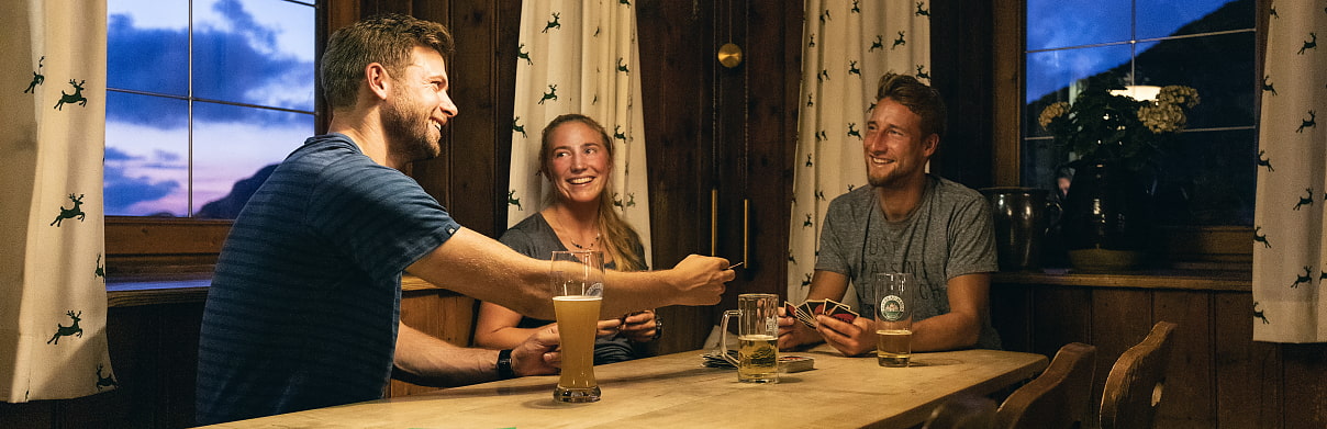 3 Wanderer sitzen gemütlich in der Stube der Lamsenjochhütte und spielen ein Gesellschaftsspiel
