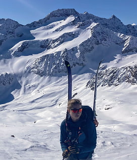 Leiterin Selina steht mit Skier am Rucksack vor einer malerischen Bergkulisse