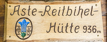 Holzschild mit der Aufschrift Aste-Reitbihel-Hütte 936m und das Logo mit blauem Enzian