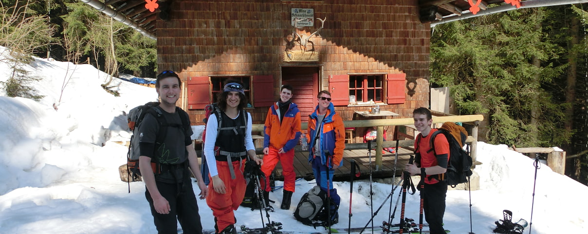 Bild zeigt 5 Skitourengeher, die gerade auf einer holzgetäfelten Hütte mit roten Fensterläden ankommen