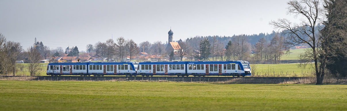 Bild zeigt den BOB Abkürzung für Bayerische Oberland Bahn in voller Länge. Im Hintergrund sieht man ein Voralpendorf mit einer Kirchen mit Zwiebelturm