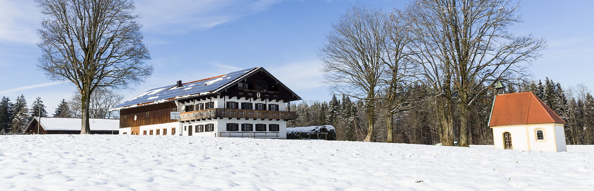 Bild zeigt den Oberländerhof Haunleiten und die kleine Kapelle in einer winterlichen schneebedeckten Landschaft