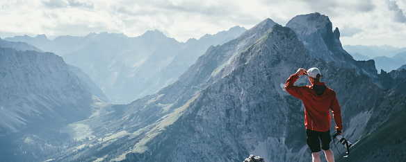 Bergwanderer mit langer roter Bergjacke blickt in die Ferne zu den Bermotiven