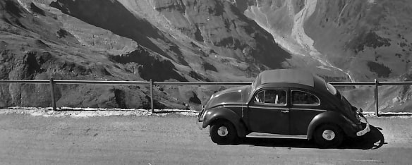 Auf dem Bild sieht man eine Schwarz-Weiß Aufnahme von einem älteren VW-Käfer auf einer Alpenstraße. Im Hintergrund ist ein alpines Gelände erkennbar. Die Straße ist durch einen Holzzaun begrenzt.  