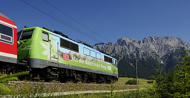 Auf dem Bild sieht man eine Lokomotive in den Farben des Alpenvereins mit der Aufschrift mit der Bahn in die Berge. Im Hintergrund sind die Berge zu sehen
