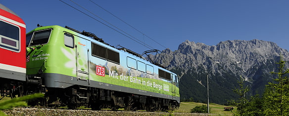 Auf dem Bild sieht man eine Lokomotive in den Farben des Alpenvereins mit der Aufschrift mit der Bahn in die Berge. Im Hintergrund sind die Berge zu sehen