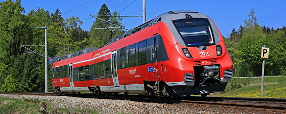 Auf dem Bild sieht man eine roten Regionalzug bestehen aus einer Lokomotive und zwei Wagons, die auf einem Gleis entlang fährt. Im Hintergrund ist eine bewaldete Landschaft und einer blauer Himmel zu sehen