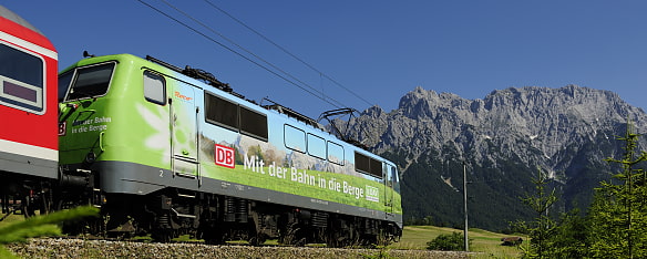 Auf dem Bild sieht man eine Lokomotive in den Farben des Alpenvereins mit der Aufschrift mit der Bahn in die Berge. Im Hintergrund sind die Berge zu sehen