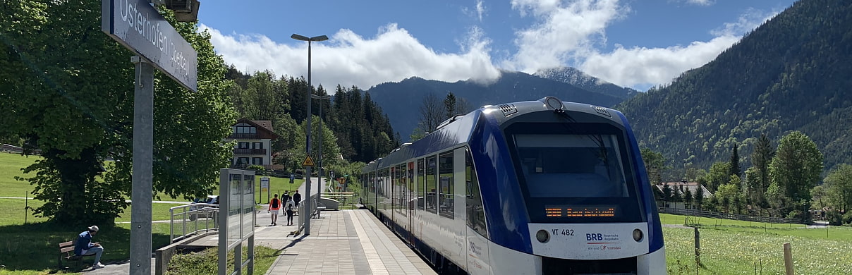 Bild zeigt eine Regionalbahn in den Farben Blau und Weiß, wie sie am Bahnhof Osterhofen steht. Im Hintergrund sind Wielsen und bewaldete Berge sowie ein blauer Himmel mit einigen weißen Wolken zu sehen. Es befinden sich auch ein paar Personen auf dem Bahnsteig.