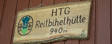 Auf dem Bild sieht man ein Holzschild mit der Aufschrift Oben in Großbuchstaben HTG und mittig darunter Reitbihelhütte und darunter mittig 940m. Rechts daneben ist in einem Kreis das blaue Edelweis und die Buchstaben A und darunter S auf der rechten Seiten und V und darunter O auf der linken Seite 