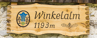 Auf dem Bild sieht man ein Holzschild mit der Aufschrift Winkelalm und darunter links bündig 1193m. Rechts daneben ist in einem Kreis das blaue Edelweis und die Buchstaben A und darunter S auf der rechten Seiten und V und darunter O auf der linken Seite 