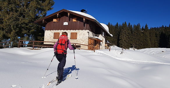 Auf dem Bild sieht man eine Person, die auf Skiern durch tiefen Schnee läuft in Richtung der Winkelmooshütte. Die Person trägt einen roten Rucksack und verwendet Skistöcke. Das Haus ist aus Stein (unterer Teil) gebaut und hat im oberen Teil eine Holzvertäfelung. Es hat ein schneebedecktes Dach. Im Hintergrund sind hohe Tannenbäume und der Himmel ist klar und blau.