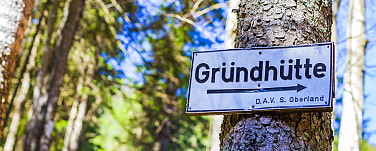 Das Bild zeigt ein weißes Blechschild an einem Baum, das  den Weg zur Gründhütte der DAV Sektion Oberland zeigt.  Es steht in schwarzer Schrift "Gründhütte" und ein schwarzer Pfeil über das ganze Schild nach rechts. Im Hintergrund sind verschwommen Bäume zu erkennen. 