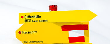 Das Bild zeigt zwei gelbe Schilder, die den Weg zur Gufferthütte und der Halserspitze zeigen. Das Schild zur Gufferthütte zeigt nach rechts. Das Schild zur Halserspitze zeigt nach links. Auf dem Schild sind auch eine Art österreichische Flagge in den Farben Rot und Weiß. Wobei der oberste Balken über die anderen beiden Balken hinausragt.