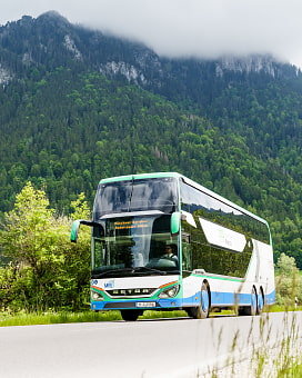 Doppeldeckerbus in den Farben weiß, blau und grün mit der Aufschrift Münchner Bergbus, der auf einer Straße in den Bergen fährt. Im Hintergrund sind bewaldete Berge zu sehen.