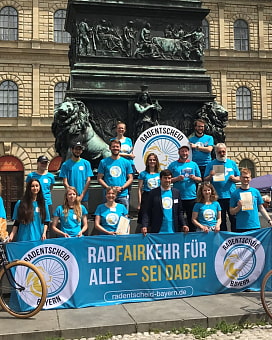 Personen mit türkisfarbenen T-Shirt, die ein Banner zum Radentscheid in der Hand halten. Sie stehen vor einem Denkmal in München.