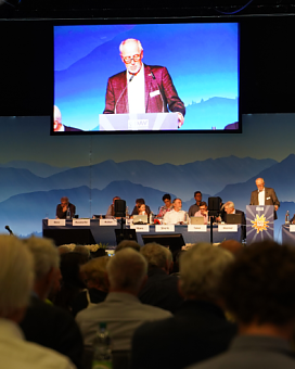 Blick vom Publikum auf das Podium der DAV-Klimakonferenz. Über dem Podium ist eine riesige Leinwand auf der der aktuelle Redner zu sehen ist, wie er an einem Pult mit Mikrofon spricht.