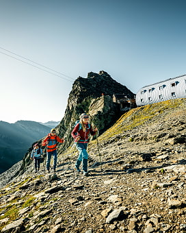 Gruppe von Bergwanderern mit Stöcken auf dem Weg nach oben zur Stüdlhütte. Im Hintergrund ist ein markanter Berggipfel und direkt daneben die Stüdlhütte. Der Himmel ist teilweise diesig und teilweise blau.