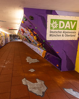 Kletterwand "Die riesige Rosi" des DAV München & Oberland in der Unterführung an der Rosenheimer Straße.  Rechts ist groß das Logo das DAV München&Oberland angebracht. Die Kletterwand ist auf der rechten Seite Violett und wird dann Flieder. Es sind viele bunte Griffe montiert.iffe
