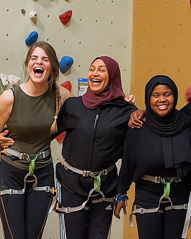 Drei Frauen verschiedener Herkunft stehen nebeneinander lachend in einer Kletterhalle