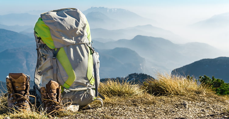 Bild zeigt einen grauen Wanderrucksack mit grüner Einarbeitung und brauen Bergschuhen die auf dem Wegrand stehen. Im Hintergrund sieht man die Berge, die leicht von Nebel verdeckt sind