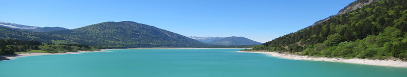 Bild zeigt einen türkisfarbigen Bergsee eingerahmt von bewaldeten Bergen. Der Himmel ist strahlend blau.
