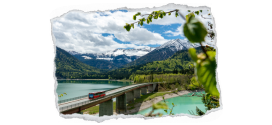 Bild zeigt einen Bus, der über eine Brücke fährt, die über einen großen See führt. Im Hintergrund die Bergwelt mit einem weiß-blauen Himmel