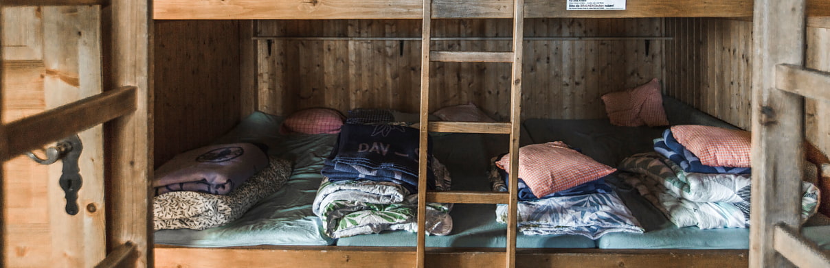 Auf dem Bild sieht man ein Schlaflager der Siglhütte. Die Betten bestehen aus mehreren Matratzen, die nebeneinander in ein großes Holzgestell gelegt sind. Jede der 4 Schlafstellen ist mit Bettwäsche, Kissen und Decken ausgestattet, die unterschiedliche Farben und Muster haben. In der Mitte befindet sich eine Holzleiter, die zu einer oberen Schlafeben oder Etage führt, die man aber auf dem Bild nicht sieht. Links im Bild sieht man noch einen Ausschnitt der Türe, die in Bettenlager führt.