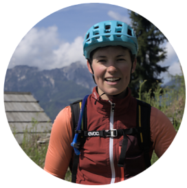 Auf dem Bild sieht man ein rundes Portrait von Katharina Filter. Sie trägt einen hellblauen Fahrradhelm und ein sportliches, rostrotes Oberteil mit Reißverschluss. Sie trägt einen schwarzen Rucksack mit einem Trinkschlauch und einer Gurtbefestigung über der Brust. Im Hintergrund befindet sich eine malerische Berglandschaft mit grünen Hängen, Nadelbäumen und einem Gebäudedach aus Holzschindeln. Der Himmel ist teils bewölkt. Es sieht so aus, als ob Sie mit dem Fahrrad unterwegs ist in den Bergen.