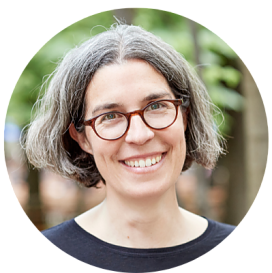 Das Bild zeigt ein rundes Portrait von Sabine Bachmann, Beiratssprecherin. Sie hat schulterlange graumelierte Haare und eine Brille mit fast runden Gläsern und einem bräunlichen Gestell. Sie lächelt ins Bild mit leicht geneigten Kopf. Der Hintergrund ist unscharf und zeigt eine Natur mit Bäumen 
