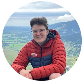 Das Bild zeigt ein rundes Portrait von Jana Wagner, Jugend & Junioren. Sie hat mittelbraune Haare, die zu einem Pferdeschwanz gebunden sind. Des weiteren trägt sie eine rahmenlose Brille und einen schwarzen Hoodie. Sie steht vor einer weißen Hauswand und schaut wahrscheinlich etwas gegen die Sonne, da sie die Augen leicht geschlossen hat.