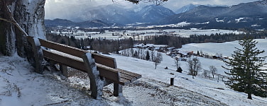 Eine schneebedeckte Aussichtsbank am Hang mit Blick ins schneebedeckte Tal