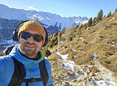 Gereon mit Kopfhörern, Mütze und Sonnenbrille. Im Hintergrund braune Wiesen und Schnee bedeckte Berge.