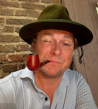 Stephan Decker augenzwinkernd mit Pfeife und Hut vor einer Backsteinwand.
