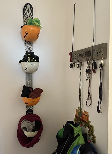 Ein Ski hängt senkrecht an der Wand. Daran befestig sind vier Kletterhelme mit der geöffneten Seite nach oben. In diesen liegen Handschuhe und Schals.