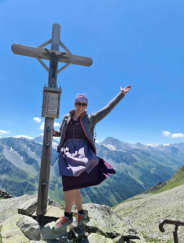 Nicole im Dirndl am Gipfelkreuz. Im Hintergrund Berge.