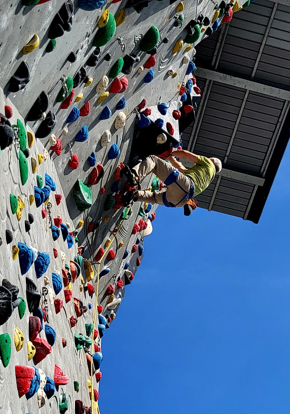 Eine kletternde Person navigiert geschickt durch eine bunte Kletterwand, gesichert durch ein Seil, vor dem Hintergrund eines strahlend blauen Himmels und eines Daches, was einen Moment sportlicher Anstrengung und Vertrauens darstellt.