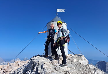 Zwei Frauen stehen mit Klettstreigset vor der Hipfelmarkierung des Triglav. 