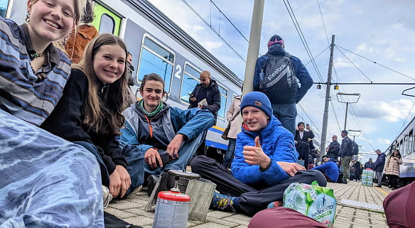 Vier Jugendliche sitzen am Bahngleis, im Hintergrund steht ein Zug. Vor den Jugendlichen steht am Boden ein Campingkocher. 
