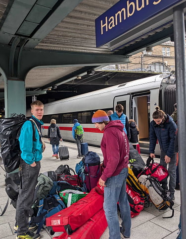 Eine Gruppe von Jugendlichen steht am Bahngleis, im Hintergrund steht ein Zug mit offener Türe. Am Boden liegt ein Haufen Gepäck mit Rücksaäcken, Skitaschen und Duffle-Bags.