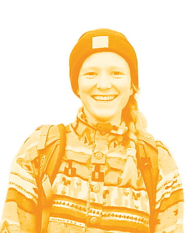 Junge Frau mit Mütze, Zopf und Rucksack lächelt in die Kamera. Sie trägt ein Hemd mit Ethnomuster.