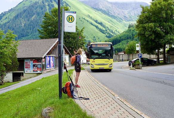 Wanderin steht an Bushaltestelle. Ein Bus fährt gerade heran. Im Hintergrund eine Schneune und Berge.