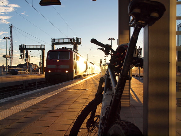 Mountainbike lehnt am Bahngleis. Ein Zug fährt gerade ein/ab. Die tiefstehende Sonne spiegelt sich in dessen Fenstern.