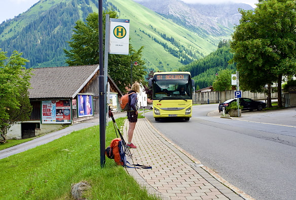 Wanderin steht an Bushaltestelle. Ein Bus fährt gerade heran. Im Hintergrund eine Schneune und Berge.