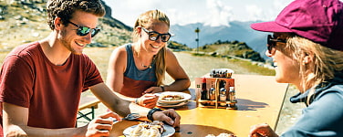 Drei Menschen sitzen an einem Tisch im Freie und essen. Im Hintergrund ist ein Bergapanorama.